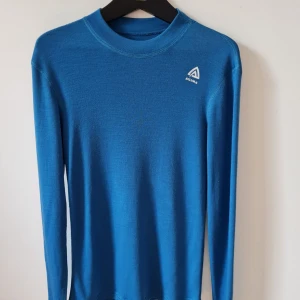 Blå långärmad tröja från Aclima strl 146 (barn) - Blå långärmad tröja från Aclima med rund hals och logga på bröstet. Tillverkad i 100% merinoull. Passformen är normal och materialet är tunt och smidigt, perfekt för lager-på-lager. Enkel och stilren design utan mönster eller detaljer. Finns små hål på nedre delen av ryggen och nedersta kanten på ryggen,  se bild. Storlek 146 cm.