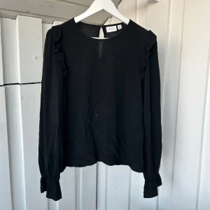 Svart blus med volang från VILA - Svart blus från VILA i storlek 36. Blusen har långa ärmar med lätt puff och volangdetaljer längs axlarna.  Tecken på att den är använd men går lätt bort i tvätten eller med vanish☺️