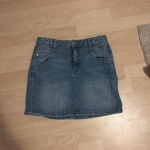 Blå jeanskjol med fickor - Säljer en klassisk blå jeanskjol med fem fickor och knappstängning fram. Kjolen har en rak passform och är i kort modell, perfekt för dig som gillar en enkel och tidlös stil. Passar till sneakers eller boots för en chill look.