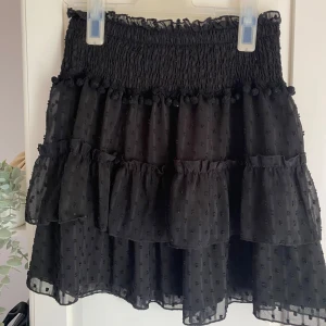 Svart volangkjol med prickar - Supersöt svart kortkjol med volanger och små prickar i tyget. Kjolen har en smockad midja och dekorativa bollar längs kanten. Perfekt för dig som gillar en lekfull och trendig stil.