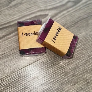 Lavendeltvål med torkad hibiskus - Handgjord tvål med lavendeldoft med torkad hibiskus i en djup lila färg, inslagen i genomskinlig plast och ett brunt pappersband med texten 'lavendel'. Perfekt för dig som älskar naturliga dofter och vill ha en snygg detalj i badrummet.