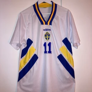 Svenska landslagströja Brolin #11 Adidas - Svensk landslagströja från Adidas med Tomas Brolin och nummer 11 på ryggen. Vit bas med blå och gula detaljer, SVFF-emblem på bröstet och klassisk v-ringad krage i blått och gult. Tillverkad i lätt och ventilerande material, perfekt för fotbollsfans.