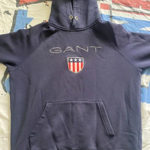 Mörkblå hoodie från GANT med logga - Säljer en mörkblå hoodie från GANT med broderad logga och sköld på bröstet. Tröjan har huva med snörning och en stor magficka. Perfekt för dig som gillar klassisk och sportig stil. Är storlek L men Passar som Storlek S/ M. 
