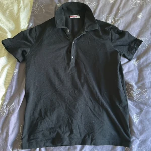 Svart kortärmad t-shirt från Prada - Svart t-shirt från Prada med klassisk krage och knappslå framtill. Tillverkad i mjuk bomull med lite stretch för extra komfort. Snygg och enkel design som funkar till många olika stilar. Perfekt för dig som gillar stilrena plagg med exklusiv känsla. Den är väl använd därav det priset 