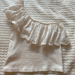 Vit oneshoulder topp med volang - Supersöt vit topp med oneshoulder och bred volangkant med broderade blommor och hålmönster. Ribbad botten i mjuk bomull och volangen i tunnare tyg. Perfekt för dig som gillar detaljer och vill sticka ut lite extra.