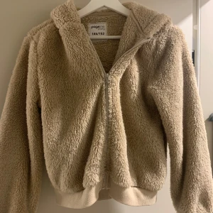 Beige teddyjacka med huva Page One - Supermjuk och fluffig teddyjacka i beige från Page One Young. Jackan har huva, dragkedja framtill och ribbade muddar vid ärmslut och nederkant. Perfekt för dig som vill ha en cozy och trendig look under kyliga dagar.