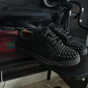 Svarta Louboutin sneakers med nitar - Snygga svarta sneakers från Louboutin med klassisk röd sula och coola svarta nitar på tån. Skorna har snörning och är tillverkade i skinn med en platt sula. Perfekta för dig som vill sticka ut med en edgy vibe.
