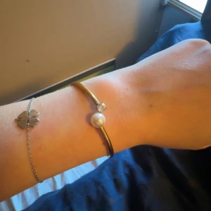 Luna Bangle Gold armband - Snyggt och stilrent guldarmband från Luna med en vit pärla och en glittrande sten på varsin ände. Armbandet är öppet och har en minimalistisk design som ger en lyxig känsla. Perfekt för dig som gillar att sticka ut med enkla men coola detaljer.