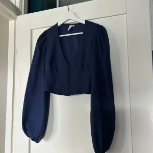 Mörkblå croppad blus från Nelly - Säljer en mörkblå croppad blus från Nelly med djup v-ringning, långa puffärmar och klädda knappar framtill. Knappt använd💕