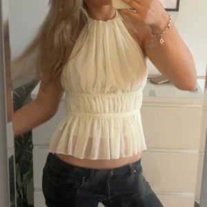 Rygglös topp - Supersöt vit blus med halterneck och volangkant nertill. Toppen har omlottkänsla och är gjord i ett tunt, luftigt material som ger en romantisk vibe. Den är lite mer krämvit än kritvit 