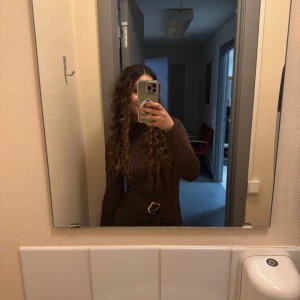 Brun långklänning med bälte - Säljer en snygg brun långklänning med lång ärm och hög hals. Klänningen har ett coolt bälte i midjan med guldfärgat spänne som ger extra stil. Passformen är slim och tyget är mjukt och stretchigt, perfekt för dig som gillar en enkel men chic look.