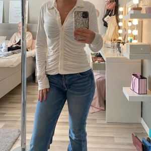 Vit långärmad skjorta med rynk - Säljer en vit långärmad skjorta med knappar framtill och snygg rynkdetalj längs hela framsidan. Skjortan har klassisk krage och figurnära passform, perfekt att styla med jeans eller kjol. 