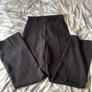 Svarta kostymbyxor i polyester M - Stilrena svarta kostymbyxor i rak modell från Kemo Moda di Zhang Xiaohua. Byxorna har hög midja, pressveck och är tillverkade i mjuk polyester med lite stretch för extra komfort. Perfekta för en clean och klassisk look.