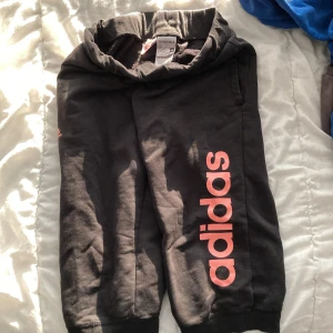 Svarta mjukisbyxor från Adidas - Svarta mjukisbyxor från Adidas med rosa logga längs benet. Byxorna har elastisk midja och muddar vid knäna för skön passform. Perfekta för chill eller träning. Storleken kan variera men det rekommenderas för barn som är 11-12 år.