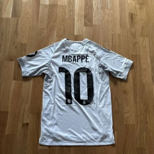 Real Madrid Mbappé 10 vit matchtröja - Säljer en officiell Real Madrid fotbollströja med Mbappé och nummer 10 på ryggen. Tröjan är vit med svarta detaljer, Adidas-logga och klubbmärke på bröstet samt Emirates Fly Better-tryck. Klassisk kortärmad modell med gula inslag längs sidorna och svarta ränder på axlarna.