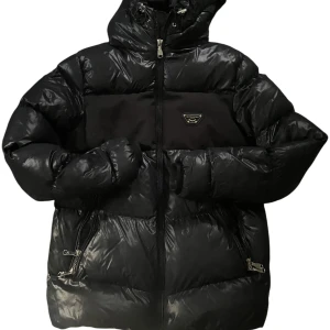 Svart pufferjacka Alessandro Milano XS - Snygg svart pufferjacka från Alessandro Milano med glansig finish och huva. Jackan har dragkedja framtill, två fickor med dragkedja och en cool logga på bröstet. Perfekt för kalla dagar när du vill hålla stilen och värmen.