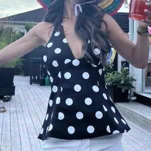 Svart prickig halterneck linnetopp - Säljer en svart halterneck topp från zara med stora vita prickar och djup v-ringning. Toppen är ärmlös och har en lätt utsvängd passform som ger en snygg siluett. Perfekt för dig som vill sticka ut med ett lekfullt mönster och cool 90-talsvibe.