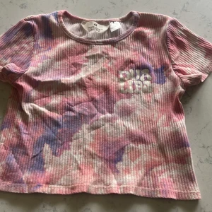 Ribbad Dua Lipa topp från H&M - Superfin ribbad topp från H&M x Dua Lipa med coolt batikmönster i rosa, lila och vitt. Kortärmad och croppad modell med tryckt Dua Lipa-logga framtill. Perfekt för dig som vill sticka ut med färg och mönster! Storlek 134-140, 20 sek + frakt