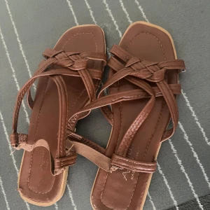 Bruna flätade sandaler med remmar - Snygga bruna sandaler med flätade remmar och fyrkantig tå. Skorna har flera tunna remmar som korsar över foten och en elastisk detalj vid hälen för extra komfort. Perfekta för varma dagar och ger en chill vibe till din outfit. Använd 2-3 gånger.