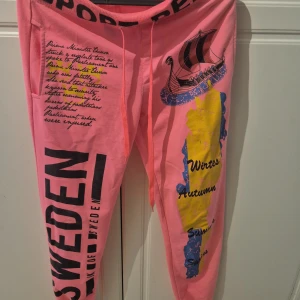 Rosa mjukisbyxor från AZN Fashion S - Unika rosa mjukisbyxor från AZN Fashion i storlek S. Byxorna har tryck med text, karta över Sverige och vikingaskepp, samt svart SWEDEN-tryck längs benet. Resår i midjan och snörning för extra komfort. Perfekta för chill dagar eller när du vill sticka ut.