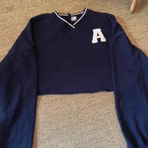 Mörkblå croppad sweatshirt med A - Mörkblå croppad sweatshirt med vit broderad bokstav A på bröstet. V-ringad hals med vita ränder och långa ärmar. Perfekt för en sportig och avslappnad stil. Mjuk bomullskänsla och loose passform.