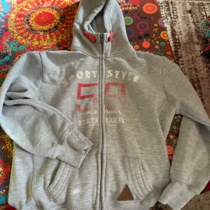 Grå hoodie med tryck och dragkedja - Grå hoodie med huva och hel dragkedja framtill. Tryck med siffran 59 och texten 'Sport Style' på bröstet. Mjukt material, två fickor fram och ribbade muddar. Perfekt för en sportig och avslappnad look.