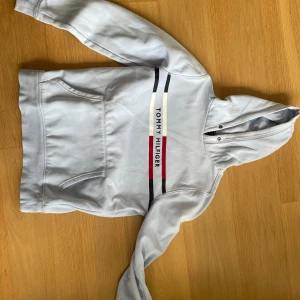 Ljusblå hoodie Tommy Hilfiger - Snygg ljusgrå hoodie från Tommy Hilfiger med klassisk logga och färgblock i vitt, rött och marinblått över bröstet. Tröjan har huva med metallöljetter, magficka och långa ärmar. Perfekt för chill eller streetwear. Materialet är mjuk bomull. Priset är ej hugget i sten
