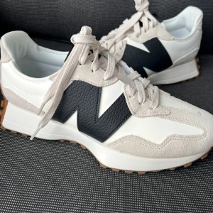 New Balance 327 vita och svarta sneakers - Snygga New Balance 327 sneakers i vitt och svart med beige detaljer. Skorna har en stor svart N-logga på sidan, ovandel i skinn och mocka samt en cool yttersula med gummidobbar. Perfekta för dig som gillar retrovibbar och streetstyle.Upplever skorna som storlek 41 därför säljer jag dem. 