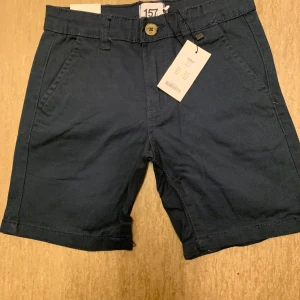Mörkblå shorts från 157, stl 130 - Säljer ett par helt nya shorts från Lager 157, modell Lee Star, i färgen mörkblå. Storleken är 130.  Shortsen är helt nya och har aldrig använts – prislappen sitter fortfarande kvar. Klassisk och stilren modell med bra passform och justerbar midja för extra komfort. Perfekta för sommaren eller till skolan!  Passar lika bra till vardag som till lite finare tillfällen. 🌟 Bekväma, snygga och helt oanvända! 