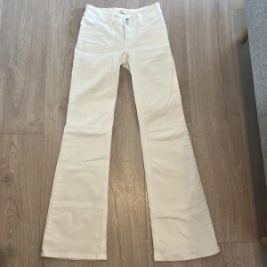 Vita lågmidjade bootcut byxor med fickor - Snygga vita bootcut-byxor med låg midja och  fickor fram samt bakfickor med knappar. Byxorna har en klassisk passform och är tillverkade i ett mjukt bomullstyg. Perfekta för dig som gillar en clean och stilren look.