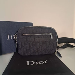Svart axelväska från Dior - Snygg svart axelväska från Dior med silverfärgade dragkedjor och diskret mönster med Dior-logga. Väskan har en justerbar axelrem och ett ytterfack med dragkedja. Perfekt för att bära det viktigaste med stil.