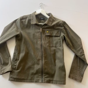 Mörkgrön overshirt från Lyle & Scott - Säljer en mörkgrön overshirt från Lyle & Scott med dragkedja framtill och klassisk krage. Jackan har en bröstficka med logotyp och två sidofickor. Jättefint skick. Passar XS-S