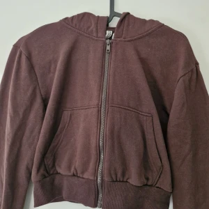 Brun croppad hoodie med dragkedja från H&M - Säljer en brun croppad hoodie med dragkedja framtill och huva. Tröjan har långa ärmar, ribbade muddar och två fickor fram. Perfekt för en avslappnad och trendig look. Noppror förekommer på ärmarna. Kan lätt tas bort med noppmaskin