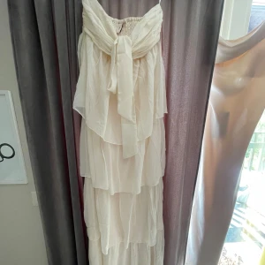 Ljus långklänning med volanger och rosett - Säljer en ljus långklänning med flera lager volanger och stor rosett framtill, perfekt för sommarens tillfällen. Aldrig använd och prislappen finns kvar💞