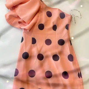 Rosa prickig klänning från Bonophard - Supersöt rosa klänning från Bonophard med stora svarta prickar och en unik volangdetalj vid axeln. Klänningen har en axelbandslös design och är perfekt för dig som vill sticka ut med ett lekfullt mönster och trendig look.