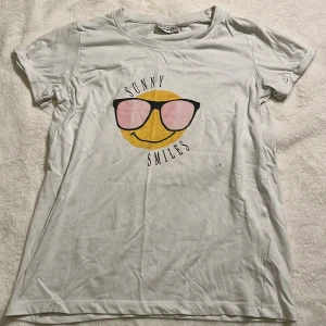 Vit t-shirt med smiley  - Vit t-shirt från Red & Blue med tryck av en gul smiley med solglasögon och texten 'Sunny Smiles' på bröstet. Klassisk rund hals och korta ärmar. Perfekt för dig som gillar en chill och lekfull stil.