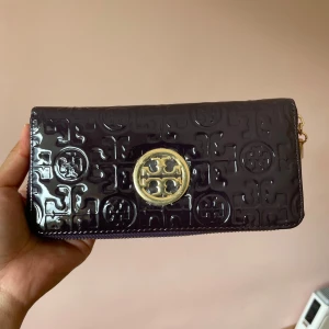 Svart plånbok från Tory Burch - Snygg svart plånbok från Tory Burch i glansigt skinn med präglat logomönster och guldfärgad logga framtill. Plånboken har dragkedja runtom och kommer med originalask och dustbag. Perfekt för dig som vill ha något stilrent men ändå lyxigt.
