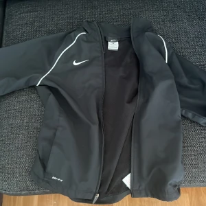 Svart träningsjacka från Nike Dri-Fit - Svart träningsjacka från Nike med vit logga och vita detaljer längs axlarna. Jackan har dragkedja framtill, meshfoder på insidan och långa ärmar med resår vid ärmsluten. Perfekt för sport och aktivitet.