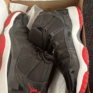 Nike Air Jordan 11 Retro 'Bred' - Säljer ett par klassiska Nike Air Jordan 11 Retro 'Bred' sneakers. Skorna är svarta med röda och vita detaljer, har hög modell och ikonisk Jumpman-logga på sidan. Ovandelen är i mesh och lackat material, med snörning framtill och rund tå.