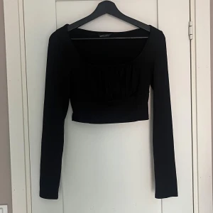 Svart croppad långärmad topp - Säljer en svart croppad topp med långa ärmar och fyrkantig ringning. Toppen har ribbat material och en rynkad detalj framtill som ger en snygg touch. Perfekt att styla med höga jeans eller kjol.