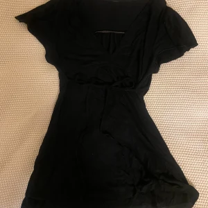 Svart kortklänning med knytband - En svart kortklänning med v-ringning och knytband i midjan. Klänningen har korta ärmar och är tillverkad i ett mjukt, lätt tyg som känns skönt mot huden. Perfekt för dig som gillar en enkel men snygg look.