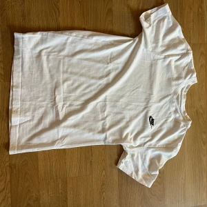 Vit Nike t-shirt med logga - Klassisk vit t-shirt från Nike med svart broderad logga på bröstet. Enkel och stilren design med rund hals och korta ärmar. Perfekt till jeans eller shorts för en clean look.