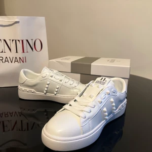 Valentino Rockstud vita  - Snygga vita Valentino Rockstud sneakers i skinn med ikoniska nitar längs sidorna och hälen. Klassisk låg modell med rund tå, platt sula och snörning. Perfekta för dig som vill ha en clean look med edgy detaljer. Kommer med originalkartong och påse.