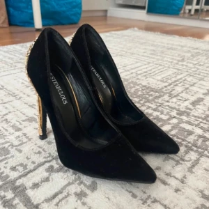 Svarta pumps med guldklack från JustFab - Svarta pumps i mocka-look med spetsig tå och höga, detaljerade klackar i guld. Klackarna har ett unikt, utsmyckat mönster som verkligen sticker ut. Perfekta för dig som vill ha något extra till outfiten.
