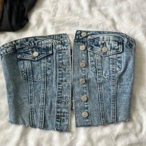 Blå jeans tubtopp med knappar - Cool tubtopp i ljusblå denim med knappar framtill och två fejkade bröstfickor. Toppen har råa kanter nertill och smockad baksida för skön passform. Perfekt för dig som vill sticka ut med en edgy jeanslook.