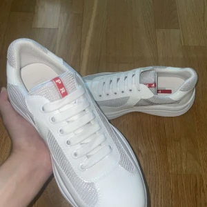 Vita sneakers från Prada - Säljer ett par fräscha vita sneakers från Prada med silvriga mesh-detaljer och klassisk röd Prada-logga på plösen. Skorna har rund tå, platt sula och snörning. Perfekta för dig som vill ha en clean och lyxig look. Passar även 38