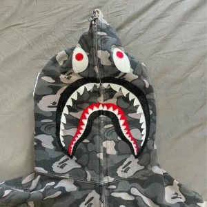 Grå camo hoodie från A Bathing Ape - Säljer en grå camo hoodie från A Bathing Ape med ikoniskt hajmotiv på huvan och broderade bokstäver. Tröjan har dragkedja hela vägen och långa ärmar. Perfekt för dig som gillar streetwear och vill sticka ut.