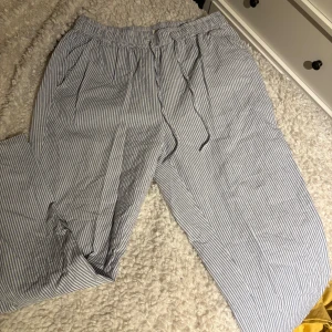 Randiga bomullsbyxor med snörning - Luftiga byxor i vit och blå randigt mönster med elastisk midja och snörning. Perfekta för varma dagar eller som chill-plagg hemma. Tillverkade i mjuk bomull för extra komfort.