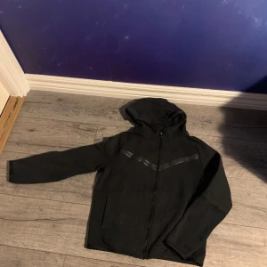 Svart hoodiejacka från Nike - Säljer en svart Nike Tech från Nike med dragkedja framtill och huva. Jackan har diskreta svarta detaljer över bröstet och är långärmad. Perfekt för en sportig och avslappnad stil.