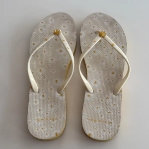 Blommiga sandaler från Tory Burch - Snygga sandaler från Tory Burch med blommigt mönster i vitt och gult. Remmarna är krämvita med små prästkragar och har en gul logodetalj. Sulan är gul på sidan och har en platt form, perfekt för varma dagar.